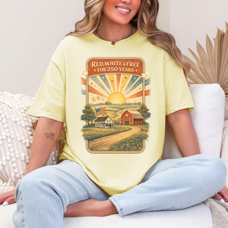 Op de afbeelding: Lichtgele t-shirt met een patriottisch ontwerp. De tekst luidt: "RED, WHITE & FREE FOR 250 YEARS." De afbeelding toont een landelijk landschap met een boerderij, schuur en Amerikaanse vlag onder een stralende zon en een kleurrijke hemel.