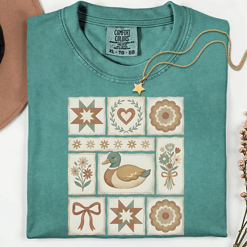May include: Teal Comfort Colors t-shirt with a patchwork design featuring a duck, flowers, stars, hearts, and geometric shapes. A gold star pendant necklace rests on the shirt. The tag reads "COMFORT COLORS XL-TG-EG".