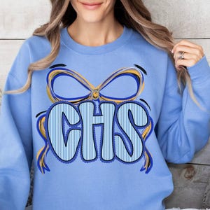 May include: A light blue sweatshirt featuring the letters "CHS" in a bold, striped font, framed by a blue and yellow bow design. The sweatshirt is a solid light blue color.