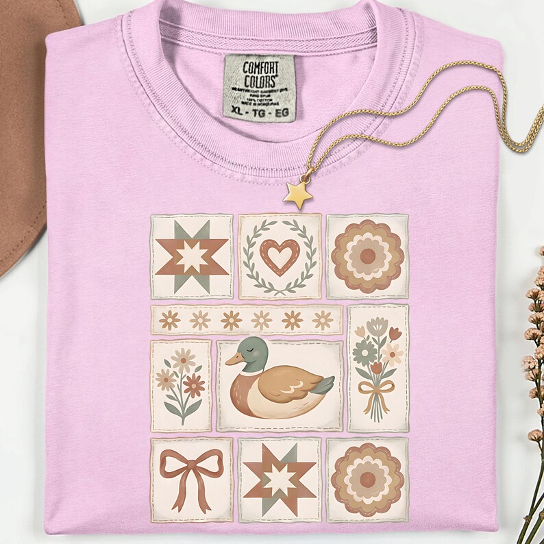 May include: A light pink t-shirt with a patchwork design featuring a duck, flowers, and geometric shapes. The shirt has a Comfort Colors tag and a gold necklace with a star pendant. The shirt is size XL.