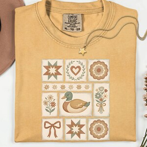 May include: Mustard yellow t-shirt with a patchwork design featuring a duck, flowers, stars, and hearts. The shirt has a Comfort Colors tag and a gold star pendant necklace. The shirt is XL.