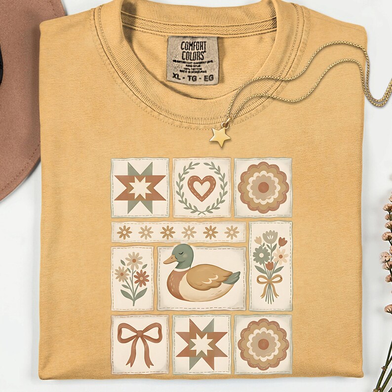 May include: Mustard yellow t-shirt with a patchwork design featuring a duck, flowers, stars, and hearts. The shirt has a Comfort Colors tag and a gold star pendant necklace. The shirt is XL.