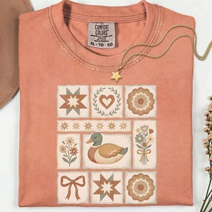 May include: Coral-colored t-shirt with a patchwork design featuring a duck, flowers, hearts, and starbursts. The shirt has a crew neck and short sleeves. A gold necklace with a star pendant is draped across the shirt. The tag reads "Comfort Colors XL-TG-EG".