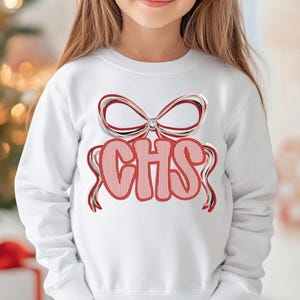 May include: White sweatshirt featuring a red and white bow design above the letters "CHS" in a red striped font. The sweatshirt has long sleeves and a crew neck. The design has a festive, celebratory feel.
