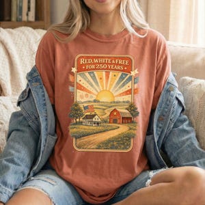 Op de afbeelding: Roestkleurig T-shirt met een patriottische afbeelding. Het ontwerp toont een zonsopgang met rode, witte en blauwe stralen, een boerderij en de tekst "Red, White & Free For 250 Years."