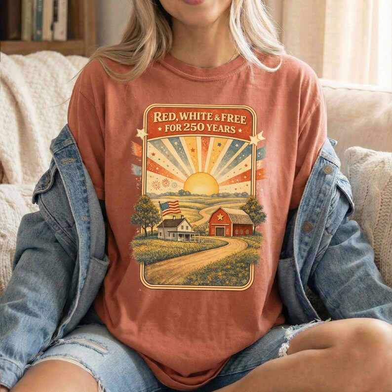 Op de afbeelding: Roestkleurig T-shirt met een patriottische afbeelding. Het ontwerp toont een zonsopgang met rode, witte en blauwe stralen, een boerderij en de tekst "Red, White & Free For 250 Years."