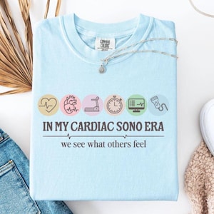 May include: Light blue t-shirt with a graphic design of a heart, a treadmill, a stopwatch, a monitor with a heart rate, and an ultrasound probe. The text on the shirt reads "In My Cardiac Sono Era We See What Others Feel."