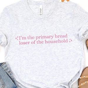 May include: A light gray heathered t-shirt featuring the pink text "I'm the primary bread loser of the household" flanked by small star icons. The shirt has a crew neckline and short sleeves, with a visible brand label inside the collar.