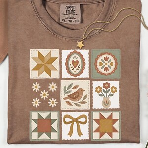 May include: A brown t-shirt with a patchwork design featuring various floral and geometric patterns in shades of beige, green, and brown. The shirt has a crew neck and short sleeves. A gold necklace with a star pendant is draped across the shirt.