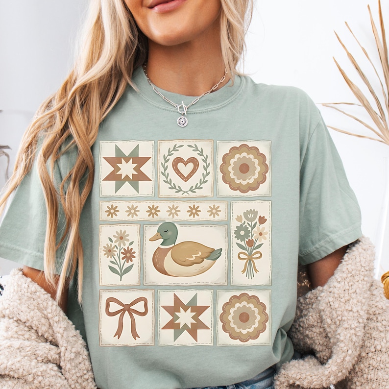 May include: A light green t-shirt featuring a patchwork design with various floral and geometric patterns. The central image is a duck. The shirt has a relaxed fit and short sleeves.
