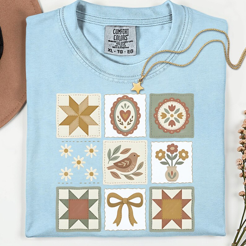 May include: Light blue Comfort Colors t-shirt with a patchwork design featuring various floral and geometric patterns. The shirt includes a tag that reads "XL - TG - EG". A gold necklace with a star pendant is draped across the shirt.