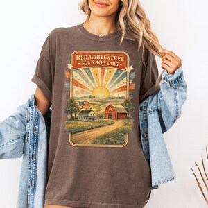 Op de afbeelding: Bruin t-shirt met een patriottische afbeelding. Het ontwerp toont een zonsopgang, een boerderij, een schuur en de tekst "RED, WHITE & FREE FOR 250 YEARS". Het shirt heeft een relaxte pasvorm.