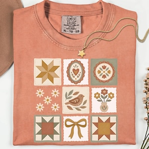 May include: A coral-colored t-shirt with a patchwork design featuring various floral and geometric patterns. The shirt has a crew neck and short sleeves. A gold necklace with a star pendant is draped across the shirt.