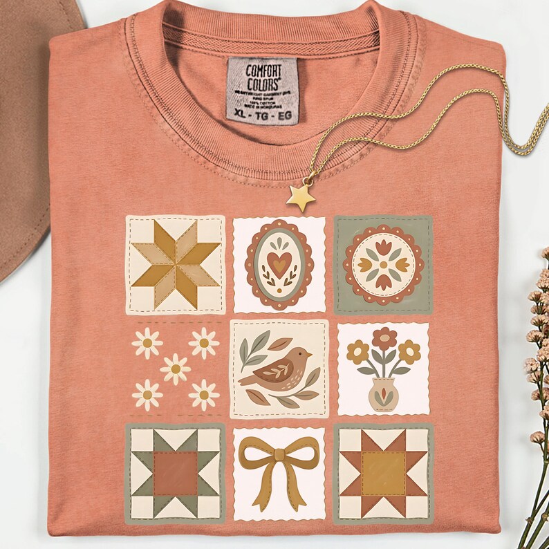 May include: A coral-colored t-shirt with a patchwork design featuring various floral and geometric patterns. The shirt has a crew neck and short sleeves. A gold necklace with a star pendant is draped across the shirt.