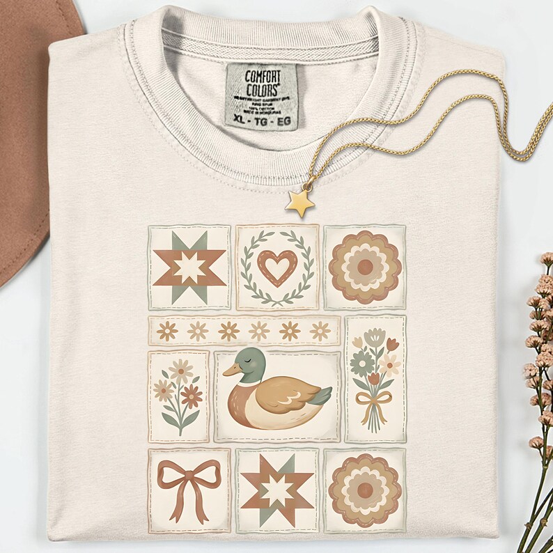 May include: Cream-colored t-shirt with a patchwork design featuring a duck, flowers, stars, and hearts. The shirt has the text "COMFORT COLORS" and size "XL - TG - EG" on the tag. A gold necklace with a star pendant is draped across the shirt.