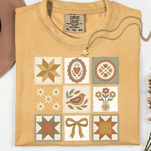 May include: Mustard yellow t-shirt with a patchwork design featuring various floral and geometric patterns in muted earth tones. A gold star pendant necklace and a Comfort Colors tag are visible. The shirt is XL.