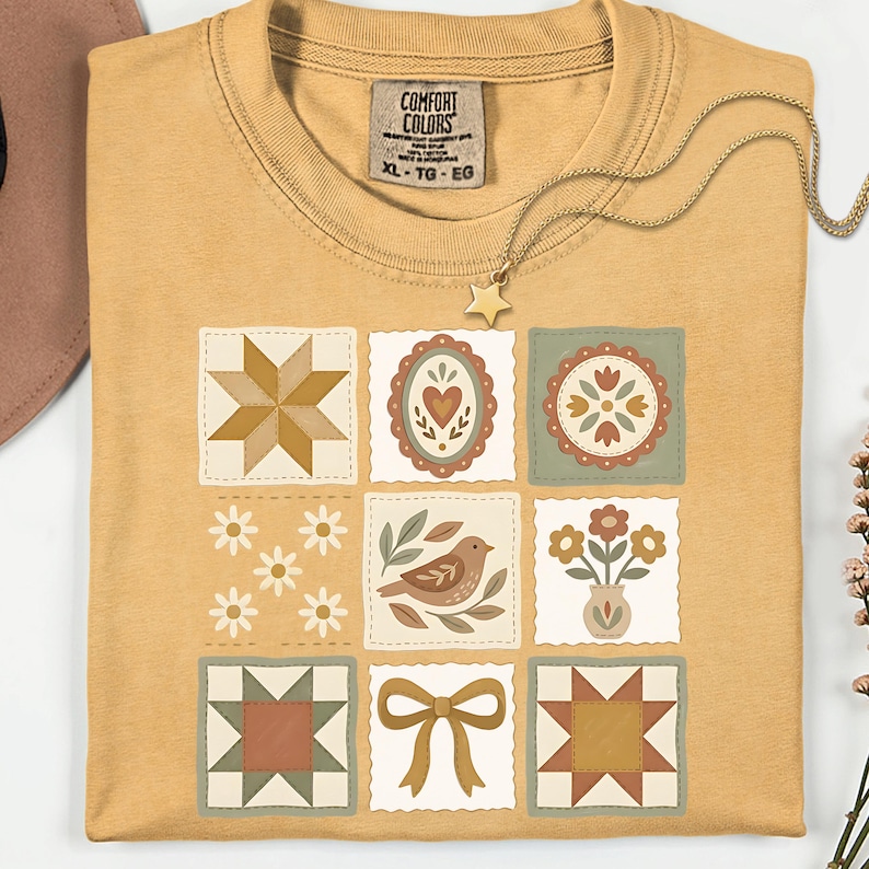 May include: Mustard yellow t-shirt with a patchwork design featuring various floral and geometric patterns in muted earth tones. A gold star pendant necklace and a Comfort Colors tag are visible. The shirt is XL.