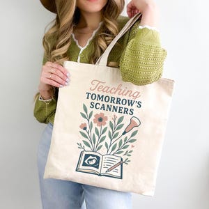 May include: Beige canvas tote bag with the text "Teaching Tomorrow's Scanners" in pink and black. The design includes floral elements, a sonogram image, and a pencil. The bag has a light-colored handle.
