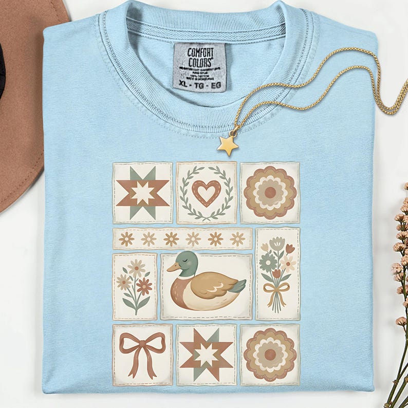 May include: Light blue t-shirt with a patchwork design featuring a duck, flowers, hearts, and starbursts. The shirt has a crew neck and short sleeves. A gold necklace with a star pendant is draped on the shirt.