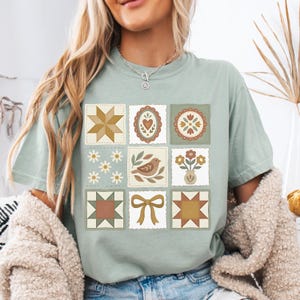May include: Sage green t-shirt featuring a patchwork design with various motifs. The design includes a star, heart, floral, bird, and bow. The squares are in shades of brown, cream, and green, creating a vintage aesthetic.