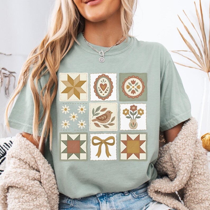 May include: Sage green t-shirt featuring a patchwork design with various motifs. The design includes a star, heart, floral, bird, and bow. The squares are in shades of brown, cream, and green, creating a vintage aesthetic.