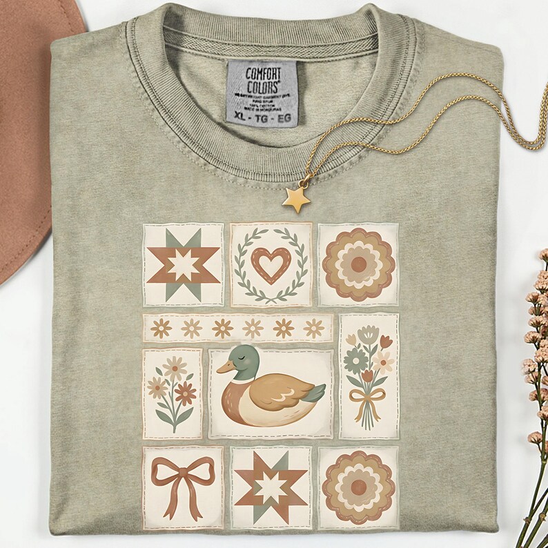 May include: A sage green t-shirt with a patchwork design featuring a duck, flowers, stars, and hearts. A gold necklace with a star pendant rests on the shirt. The shirt has a Comfort Colors tag, size XL-TG-EG.