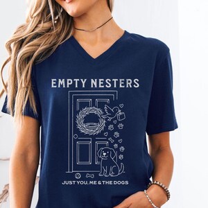 May include: Navy blue V-neck t-shirt with the text "EMPTY NESTERS" and "JUST YOU, ME & THE DOGS". The graphic features a door with a wreath, bird, dog, paw prints, and bones. The shirt is made of a soft material.