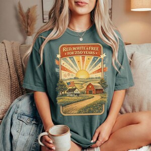Op de afbeelding: Tealkleurig T-shirt met een patriottische afbeelding. De afbeelding toont een landelijk landschap met een boerderij, schuur en Amerikaanse vlag, met de tekst "RED, WHITE & FREE FOR 250 YEARS" in een rechthoekig kader.