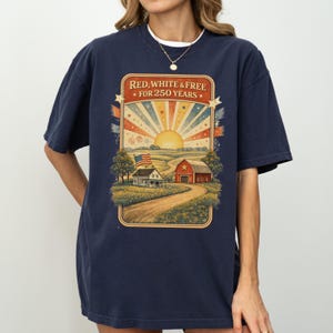 Op de afbeelding: Marineblauw T-shirt met een vintage afbeelding. De afbeelding toont een landelijk landschap met een boerderij, schuur en de Amerikaanse vlag, met de tekst "RED, WHITE & FREE - FOR 250 YEARS" bovenaan.