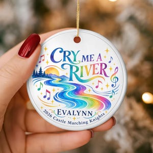 May include: A round white ceramic ornament featuring a colorful rainbow river graphic, a yellow sun, and blue pine trees. The text reads "CRY ME A RIVER", "EVALYNN", and "2026 Castle Marching Knights". Musical notes and stars surround the design.