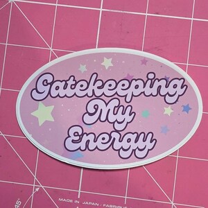 May include: Oval sticker with the text "Gatekeeping My Energy" in a purple, bubble-style font. The sticker has a pink background with pastel stars. The sticker is on a pink cutting mat.