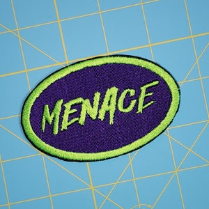 May include: A purple oval patch with a green border and the word "MENACE" embroidered in green.