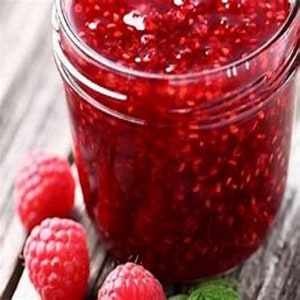 May include: A glass jar filled with red raspberry jam. The jam is thick and has a smooth texture. There are fresh raspberries on the table next to the jar.