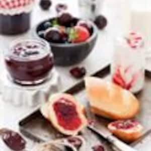 May include: A close-up of a glass jar filled with dark red homemade jam, with a spoon resting on top. The jam is spread on a small white bread roll, and there are fresh blackberries and strawberries in a bowl nearby.