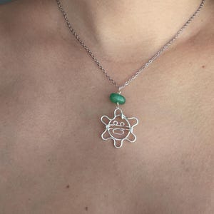 May include: Silver-toned wire pendant necklace featuring a turtle design. The pendant is suspended from a delicate chain and includes a green bead. The turtle has a smiling face.