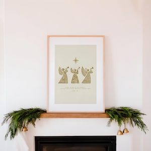 May include: A framed print featuring three golden angel figures playing trumpets beneath a star. The print has text at the bottom. The frame is light-colored, and the print is displayed above a fireplace decorated with greenery and bells.