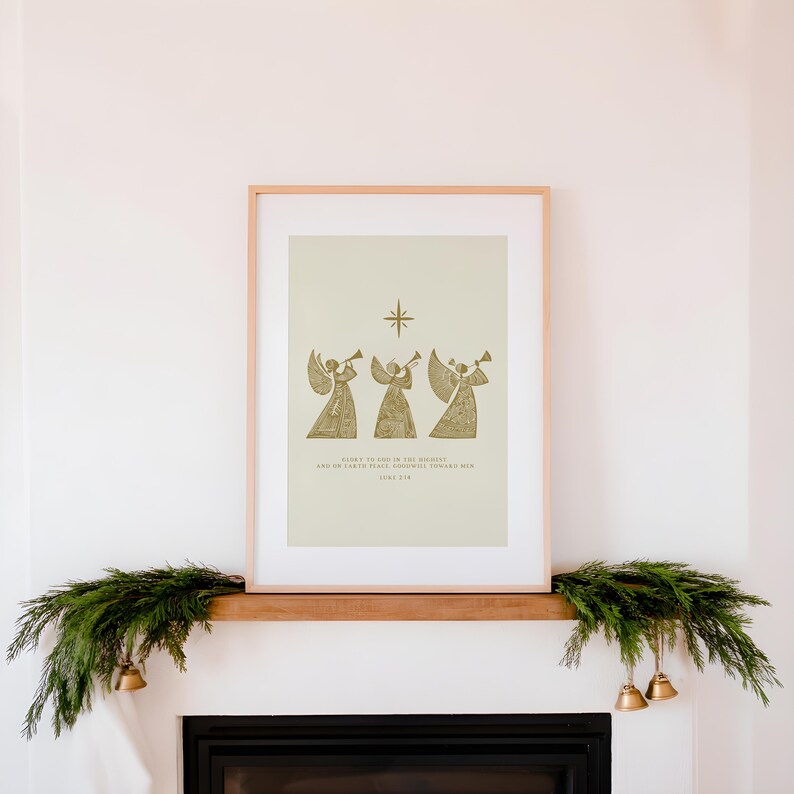 May include: A framed print featuring three golden angel figures playing trumpets beneath a star. The print has text at the bottom. The frame is light-colored, and the print is displayed above a fireplace decorated with greenery and bells.