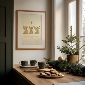 May include: A framed print with a gold illustration of three angels playing trumpets, with text below. Three green patterned mugs sit on a wooden tray, with a plate of cookies and a small Christmas tree in the background.