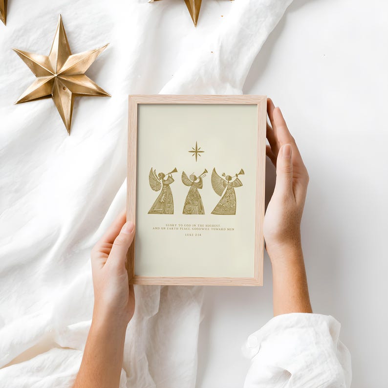May include: A framed print featuring three golden angels playing trumpets, with a star above them. The print has text at the bottom. The frame is light wood. The image is held by hands against a white background, with gold star decorations.