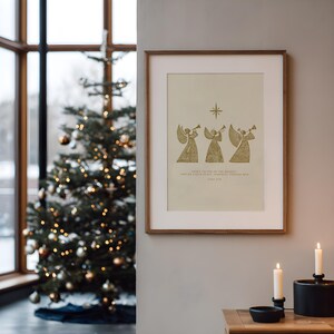May include: A framed print with a gold illustration of three angels playing trumpets, a star, and text. The print is in a wooden frame and hangs on a gray wall. A Christmas tree with lights and ornaments is in the background.