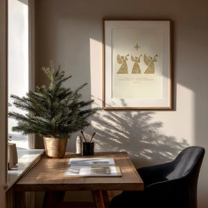 May include: A framed print with a gold illustration of three angels playing trumpets, with text below. A small Christmas tree in a gold pot sits on a wooden desk. A black chair is next to the desk, and a window is to the left.
