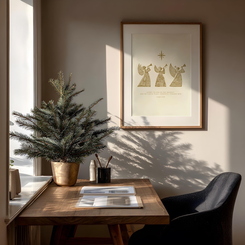 May include: A framed print with a gold illustration of three angels playing trumpets, with text below. A small Christmas tree in a gold pot sits on a wooden desk. A black chair is next to the desk, and a window is to the left.