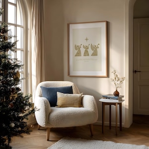 May include: A cozy living room scene with a Christmas tree, a cream-colored armchair, and a framed print. The print features three golden angel figures playing trumpets. Two pillows in blue and tan rest on the chair. A small side table holds books and a vase.