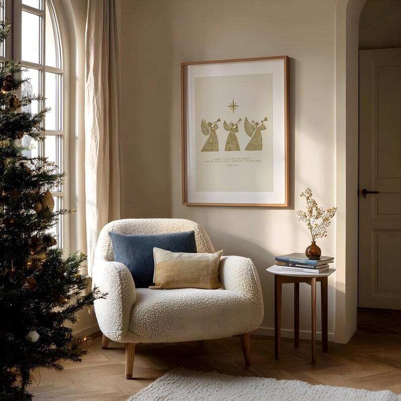 May include: A cozy living room scene with a Christmas tree, a cream-colored armchair, and a framed print. The print features three golden angel figures playing trumpets. Two pillows in blue and tan rest on the chair. A small side table holds books and a vase.