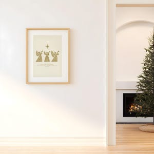 May include: A framed print featuring three golden angel figures playing trumpets beneath a star. The artwork has a light beige background and is set in a light wood frame. The print is hanging on a white wall.