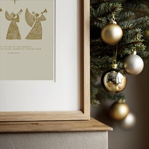 May include: A framed print with gold angel illustrations and text from Luke 2:14. The print is displayed next to a Christmas tree adorned with gold and silver ornaments. The frame is made of light-colored wood.