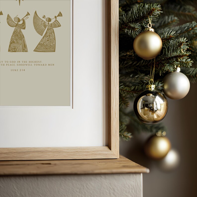 May include: A framed print with gold angel illustrations and text from Luke 2:14. The print is displayed next to a Christmas tree adorned with gold and silver ornaments. The frame is made of light-colored wood.