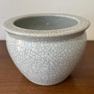 Puede incluir: Jardinera redonda de cerámica gris claro con acabado craquelado. La jardinera tiene un borde ancho y ligeramente ensanchado y un cuerpo redondeado. El interior es de un tono gris más oscuro. Adecuado para plantas pequeñas o como acento decorativo.