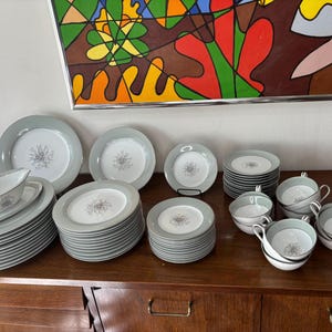 Może przedstawiać: Kompletny zestaw zabytkowej zastawy stołowej w miękkiej szaro-białej kolorystyce. Zestaw zawiera talerze, miski, filiżanki i sosjerkę, wszystkie z delikatnym kwiatowym wzorem. Naczynia są starannie ułożone na drewnianej powierzchni.