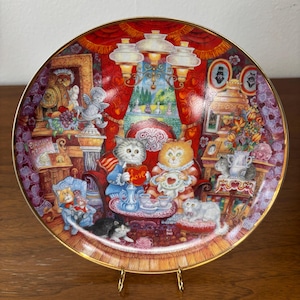 May include: Decorative plate featuring a whimsical scene of cats in a Victorian-style setting. The plate has a gold rim and depicts cats dressed in human clothing, gathered around a table with teacups. The background is a rich red with ornate details.